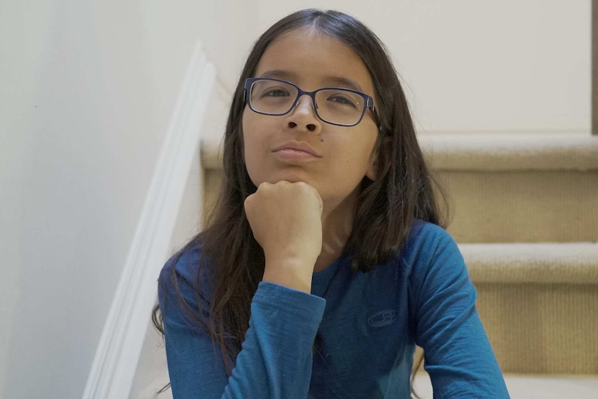 12 year old boy with long brown hair, glasses, blue t-shirt sitting on stairs looking at camera with hand on his chin