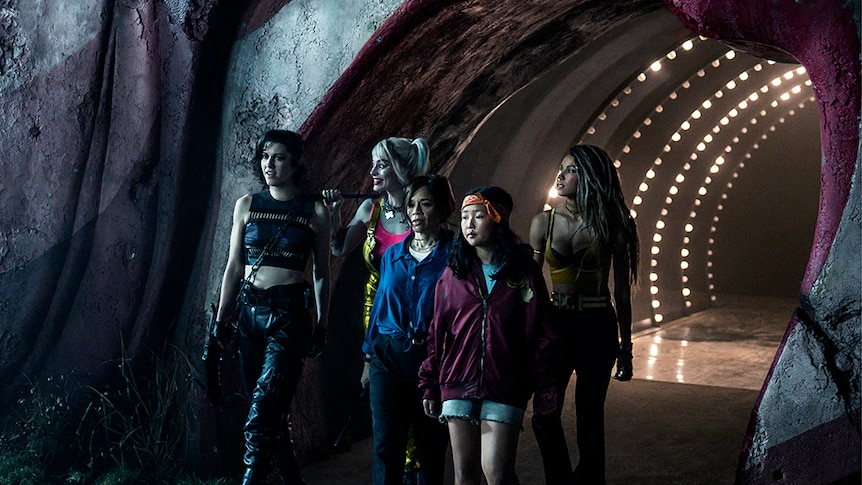 A group of five women walk through a underground tunnel, behind them a tunnel light bulb lined arched ceiling.