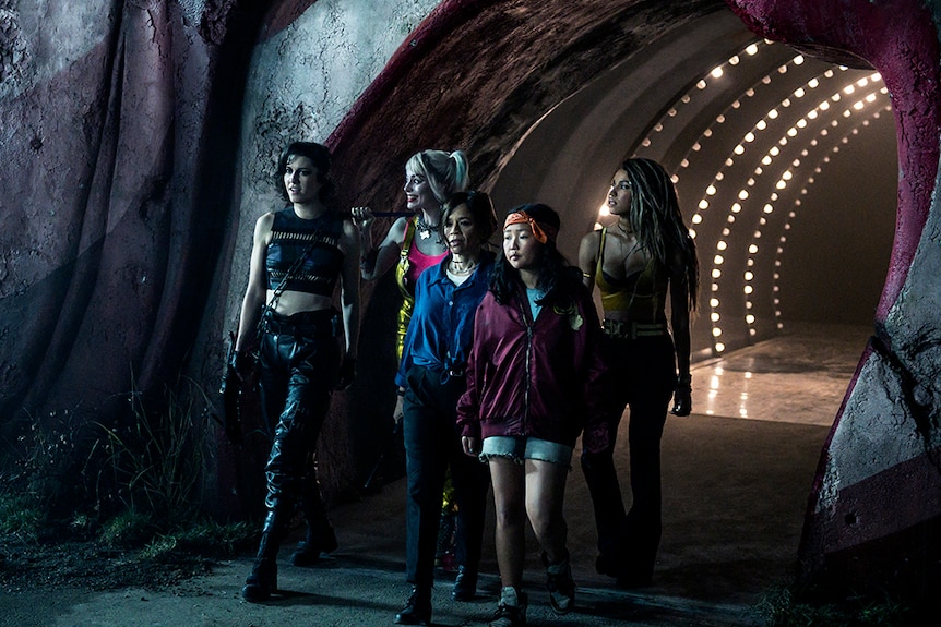 A group of five women walk through a underground tunnel, behind them a tunnel light bulb lined arched ceiling.
