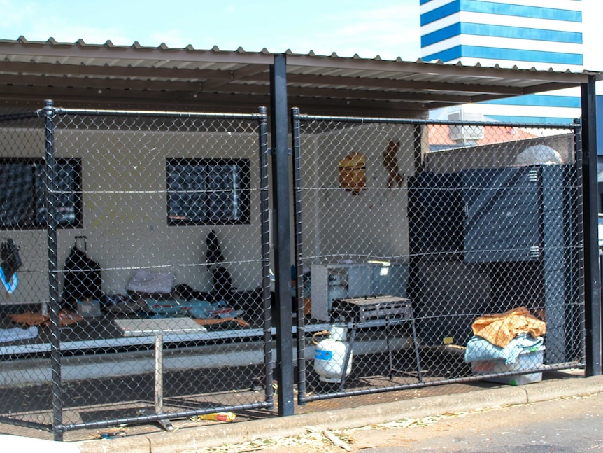 An open donga with a fence around it with beds and a gas bbq. The dongas sit within a carpark with a building behind it.