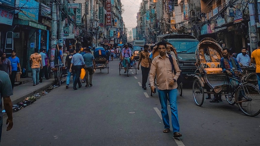 A bustling streetscape containing rickshaws and cars, with ageing buildings to either side