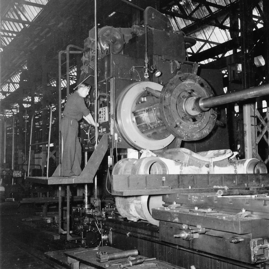 An old black and white image of a man on a platform working on a train with large wheels pictured in front of him.