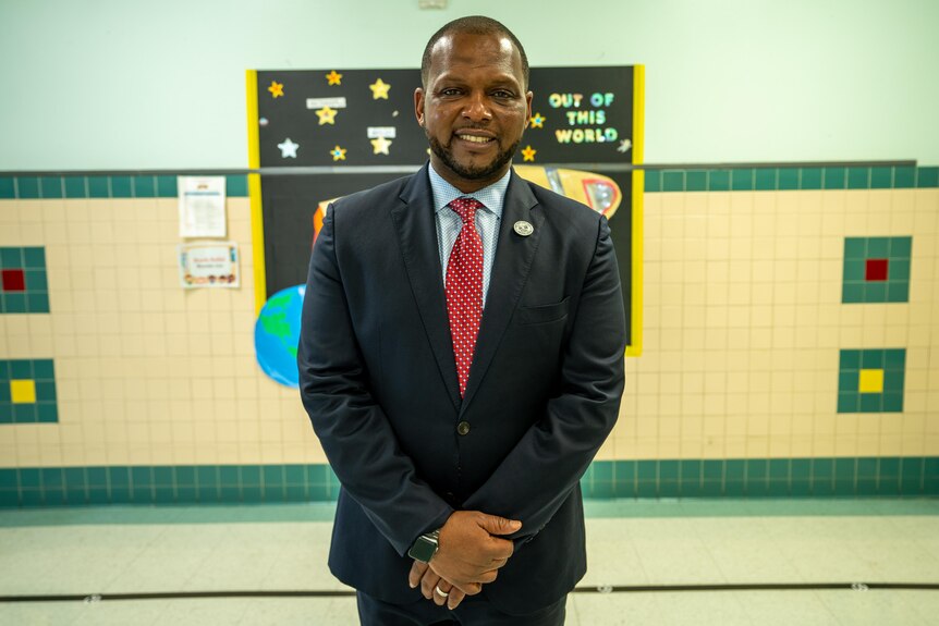 A man in a suit stands smiling in a school hallway 