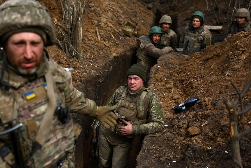 Six soldiers in camoflague fatigues stand in a narrow, muddy, chest-high trench.