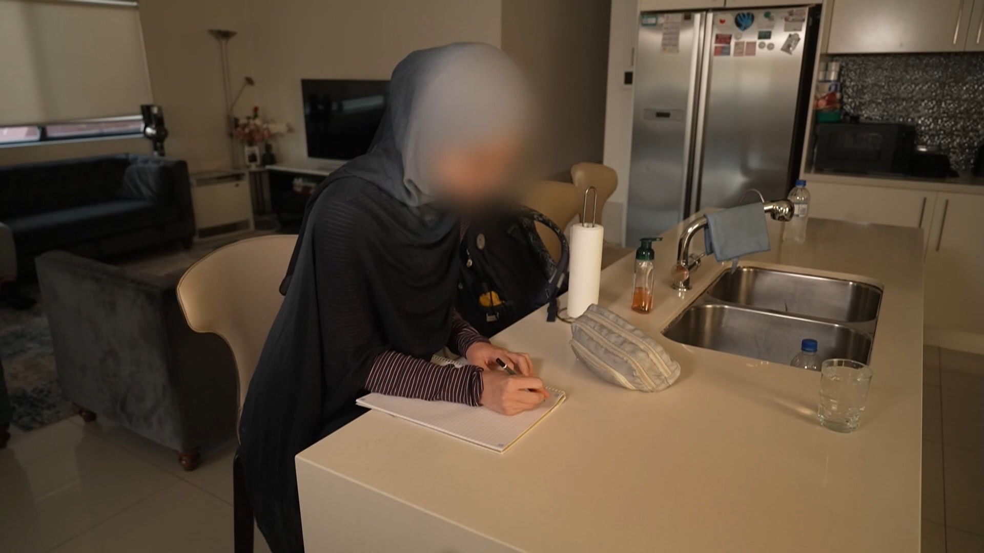 Janna, wearing a hijab, sitting at a kitchen bench writing in a note pad.