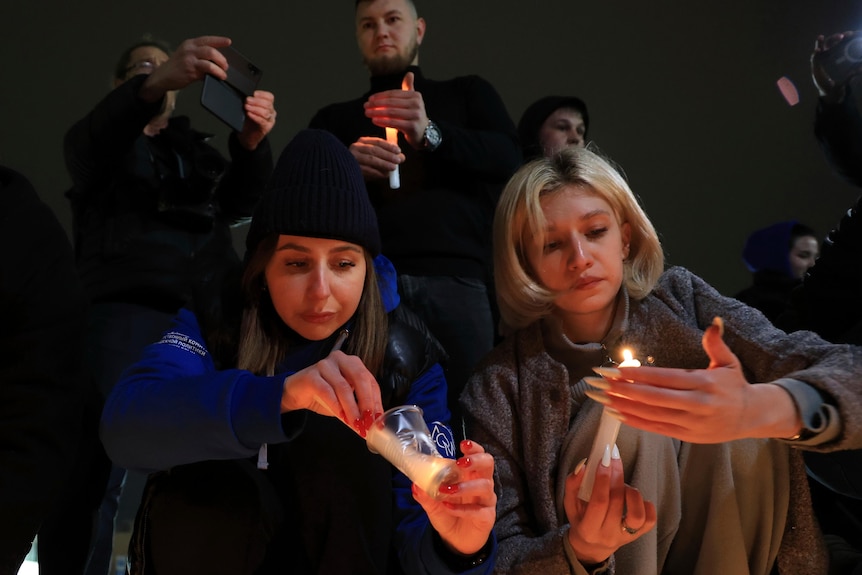 Two women crouch down to light candles in a crowd of people at a vigil