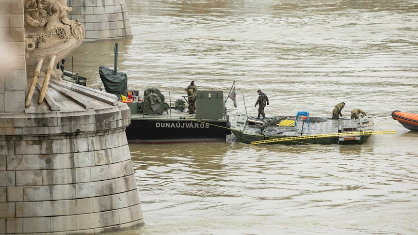 Military men stand on a vessel at the base of a bridge on the water.