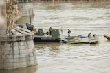 Military men stand on a vessel at the base of a bridge on the water.