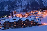 A shot from high up on a snowy mountain looks down towards a ski village in the evening, with chalets lit up and empty slopes