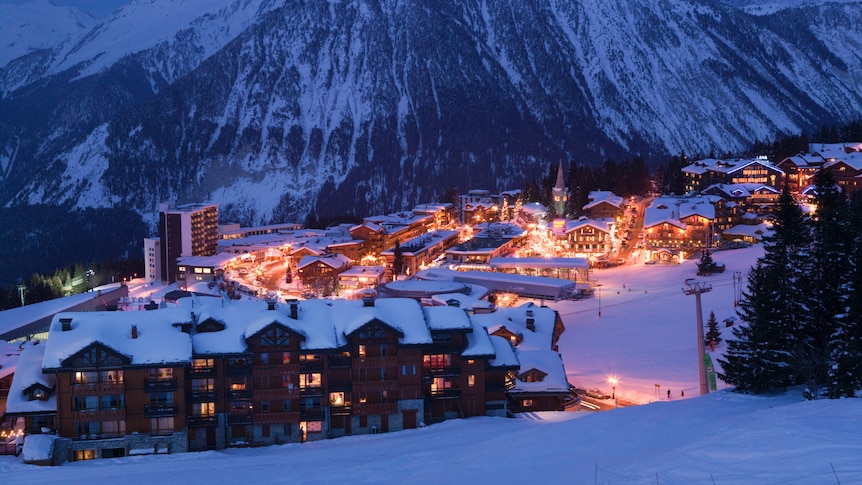 A shot from high up on a snowy mountain looks down towards a ski village in the evening, with chalets lit up and empty slopes