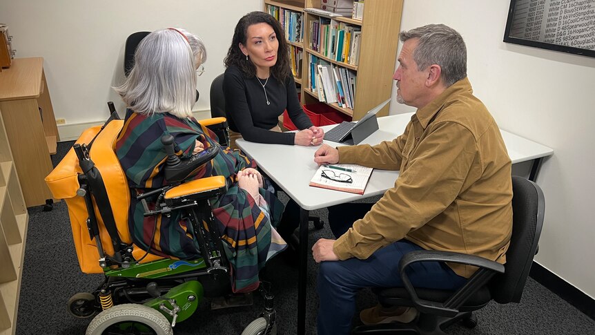 Three middle aged white people sit around a table in an office, one is in a wheelchair, the others are on office chairs.
