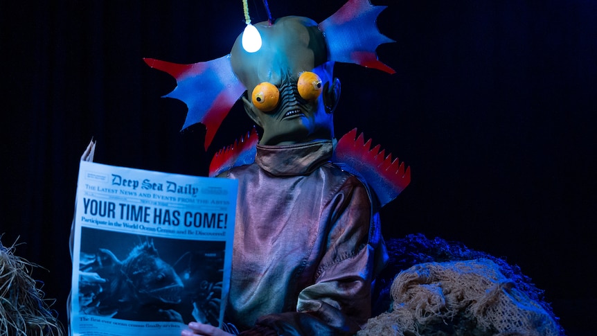 Someone dressed in a deep sea fish costume reading a pretend newspaper with the title 'Your time has come!'.