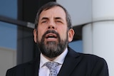 A headshot of a man with a beard wearing a suit in front of an office building.