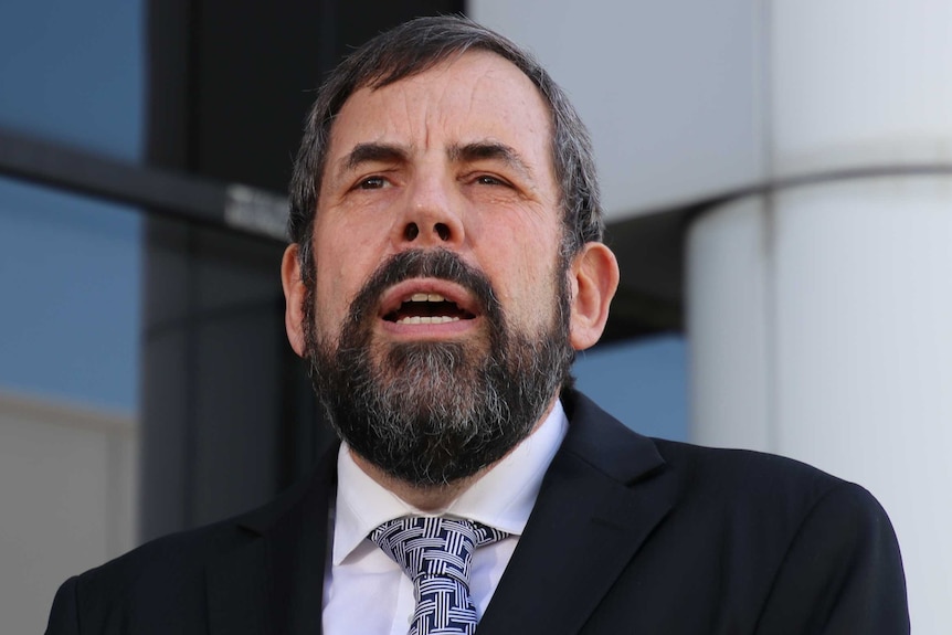 A headshot of a man with a beard wearing a suit in front of an office building.