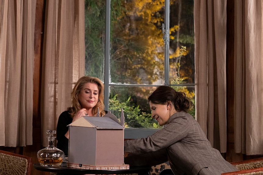 A blonde and brunette woman sit with small constructed house in living room at night time.