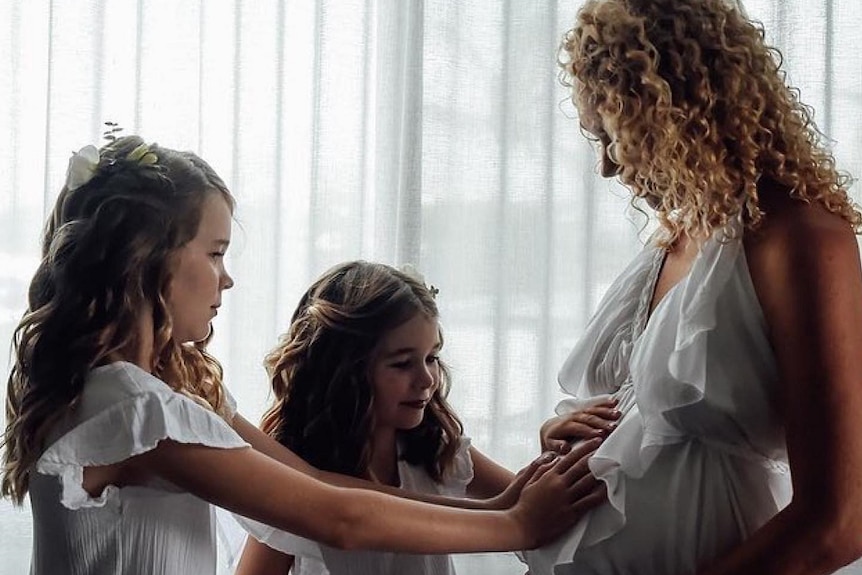Two girls in dress pictured touching their pregnant mother's belly