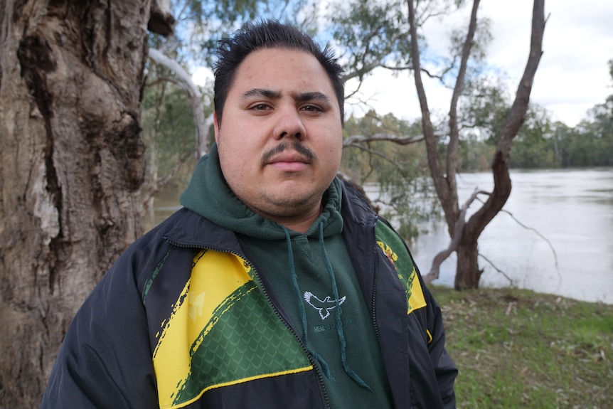 A serious looking man standing in front of a river, slight moustache, green and yellow jacket.