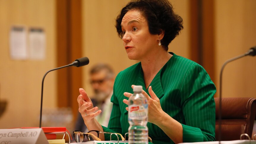 A woman with short curly brown hair gestures as she speaks. She is wearing a bright, deep green collared shirt.