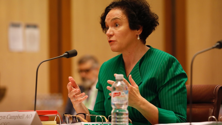 A woman with short curly brown hair gestures as she speaks. She is wearing a bright, deep green collared shirt.