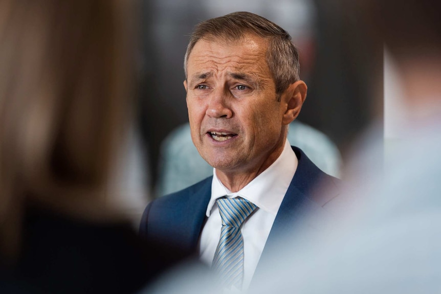 A head and shoulders shot of WA Health Minister Roger Cook speaking in a suit with out-of-focus reporters in the foreground.