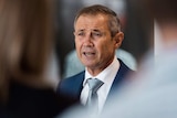 A head and shoulders shot of WA Health Minister Roger Cook speaking in a suit with out-of-focus reporters in the foreground.