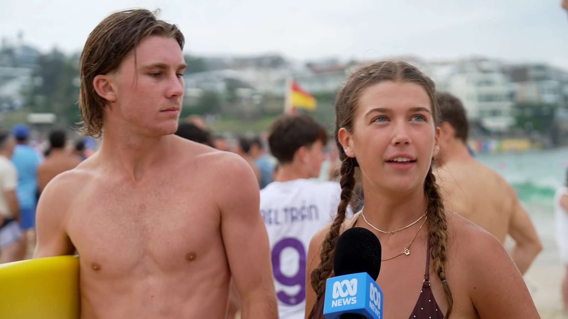 Um homem segurando uma prancha de surf amarela está ao lado de uma mulher com cabelos trançados falando em um microfone ABC.