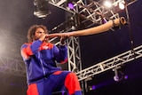 Indigenous man with curly brown hair wears blue and red tracksuit and plays a didgeridoo on stage.