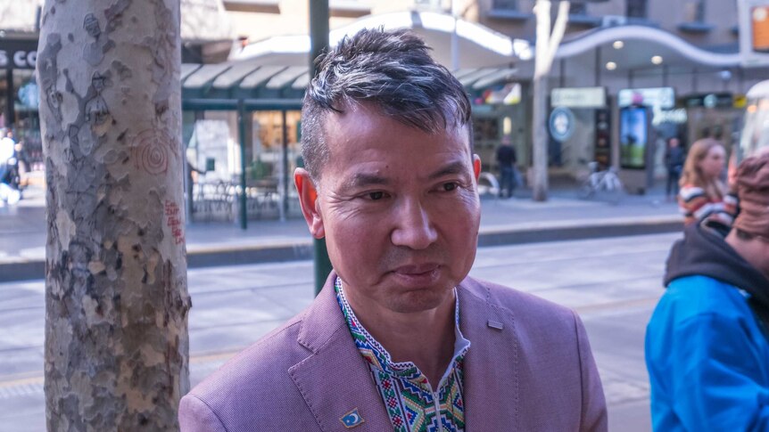 A Uyghur man wearing a traditional colourful stitched shirt and pink blazer with a Uyghur flag pin.