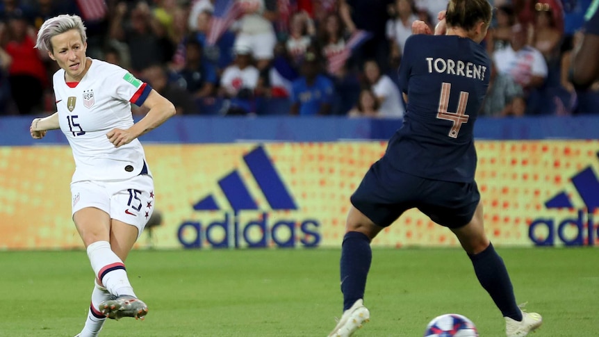 Megan Rapinoe side foots a ball through the legs of a French opponent wearing dark blue kit