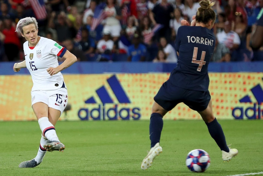 Megan Rapinoe side foots a ball through the legs of a French opponent wearing dark blue kit