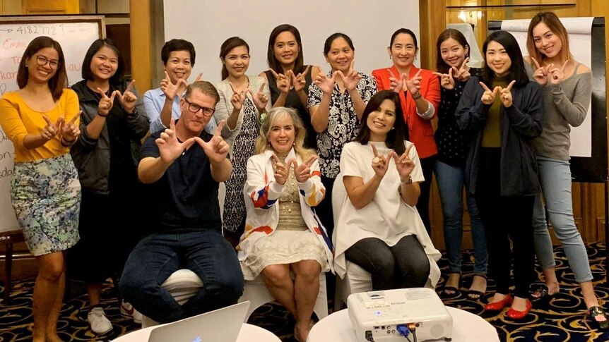 A group of women in a room stand together in the back row with three people in the front row - making a W sign with their hands