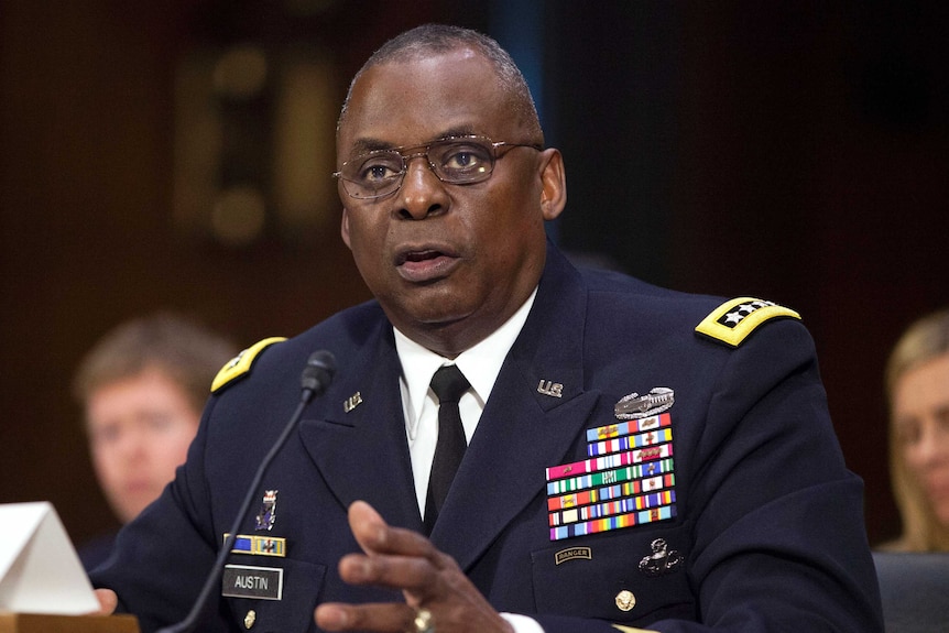 Retired General Lloyd Austin sits at a table behind a name plate wearing a jacket with badges