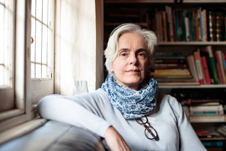 The writer, an older woman, poses in front of a bookshelf