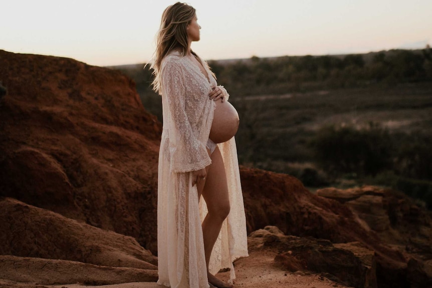 A woman wearing a lacy robe with a bare pregnant belly stands on cliffs overlooking a floodplain with soft lighting.
