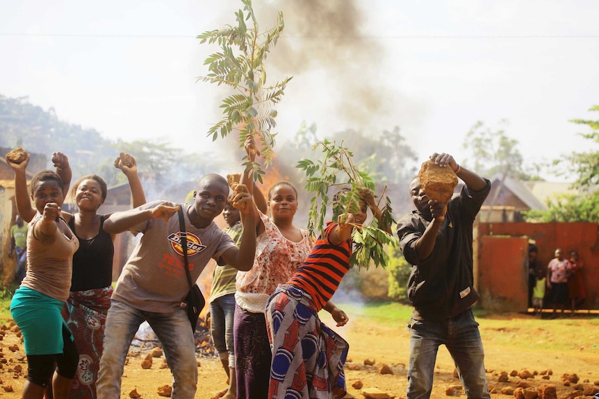 Seven people in a line-up strike different poses, one holding a tree branch, as a barricade burns behind them.
