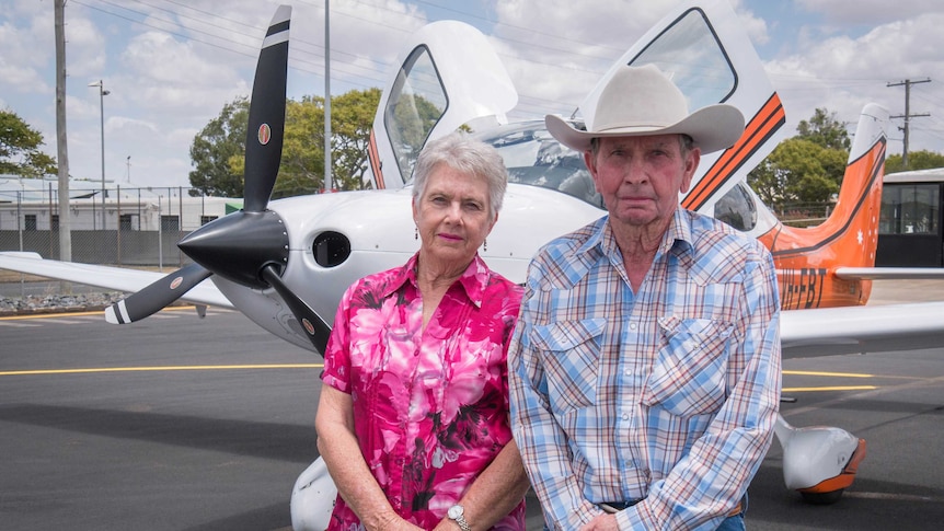 A man and a woman stand in front of a small plane, the woman wears a pink button up shirt and the man wears check and a big hat