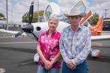 A man and a woman stand in front of a small plane, the woman wears a pink button up shirt and the man wears check and a big hat