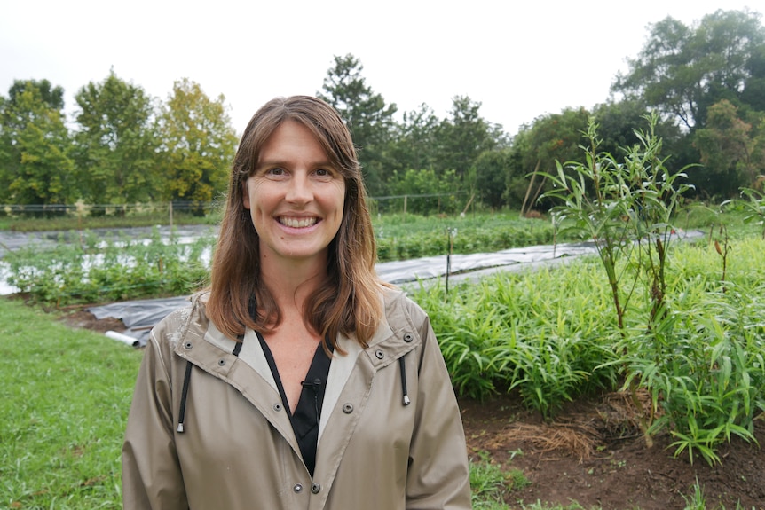 Rebecca stands in front of a ginger crop, wearing a brown raincoat, smiling.