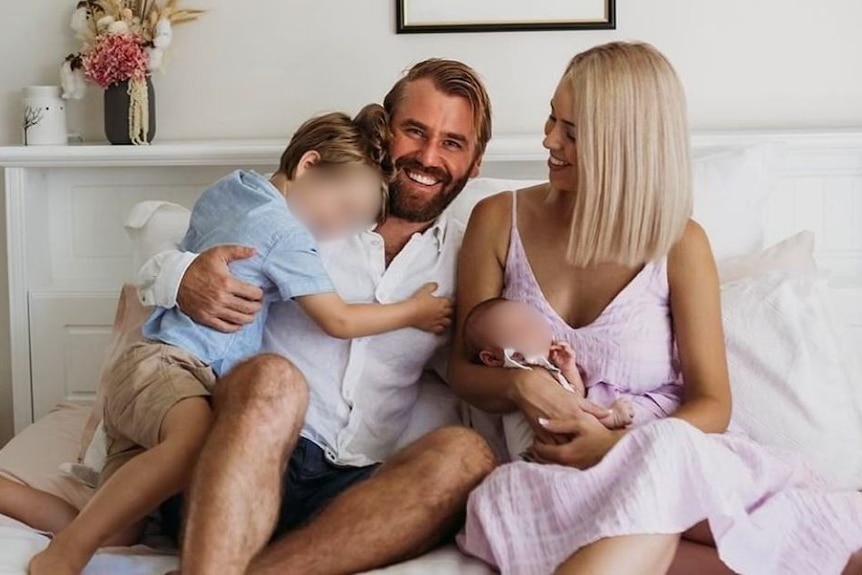 Семейное фото четырех человек: Дэниел, двое его детей и его жена Кейтленд улыбаются на белой кровати.
