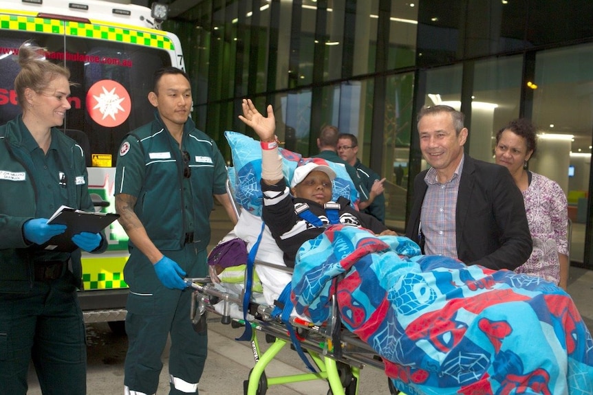 A young boy waves while lying on a stretcher at the back of an ambulance with paramedics and the WA Health Minister nearby.