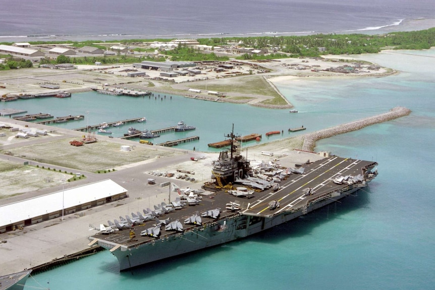 An aerial photo shows a large US aircraft carrier moored next to a base sitting on a slither of land in tropical waters.