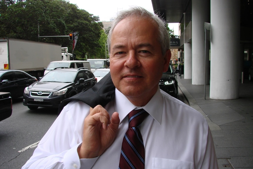 An older man in business attire, with a coat slung over his shoulder, walks down a city street.