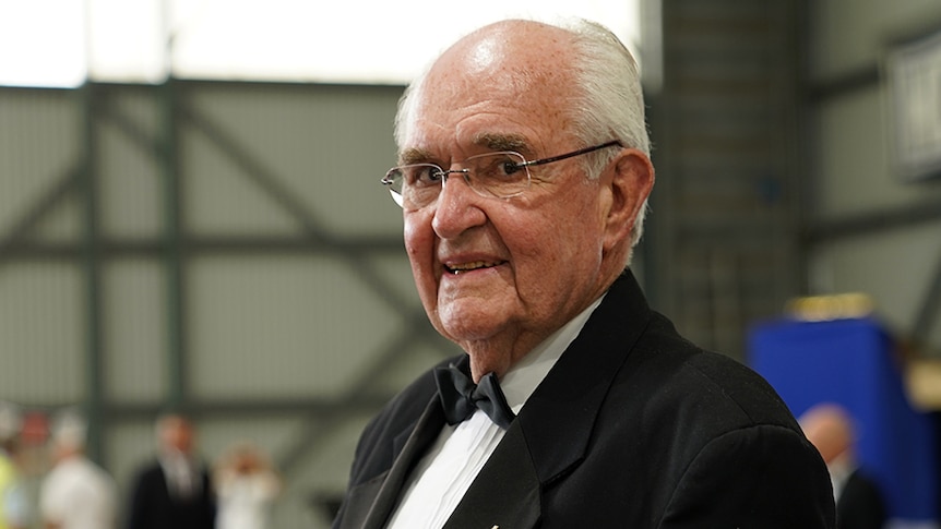 An elderly man smiles inside an airport hangar wearing a black suit and bow tie.