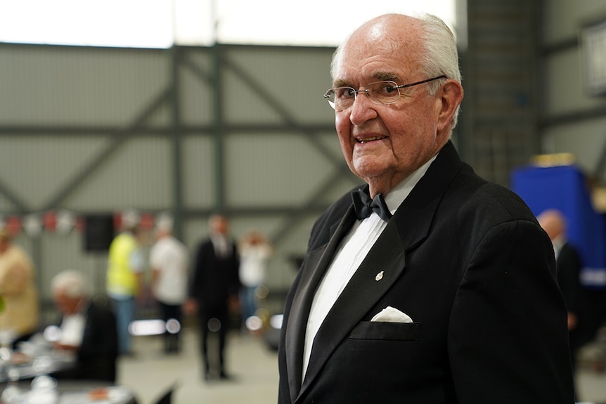An elderly man smiles inside an airport hangar wearing a black suit and bow tie.