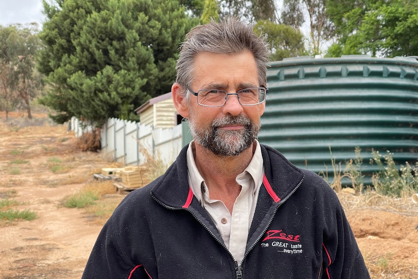 A white man, Jason Size, with glasses grey hair and beard stands in front of a green water tank on a rural property. 