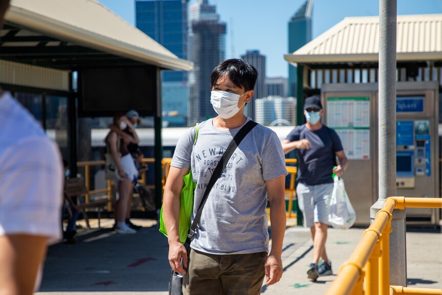 A man wearing a face mask walks away from the ferry terminal in South Perth with the CBD and Swan River behind them.