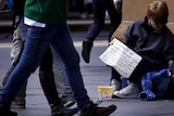 A woman holds a sign asking for money as pedestrians walk past