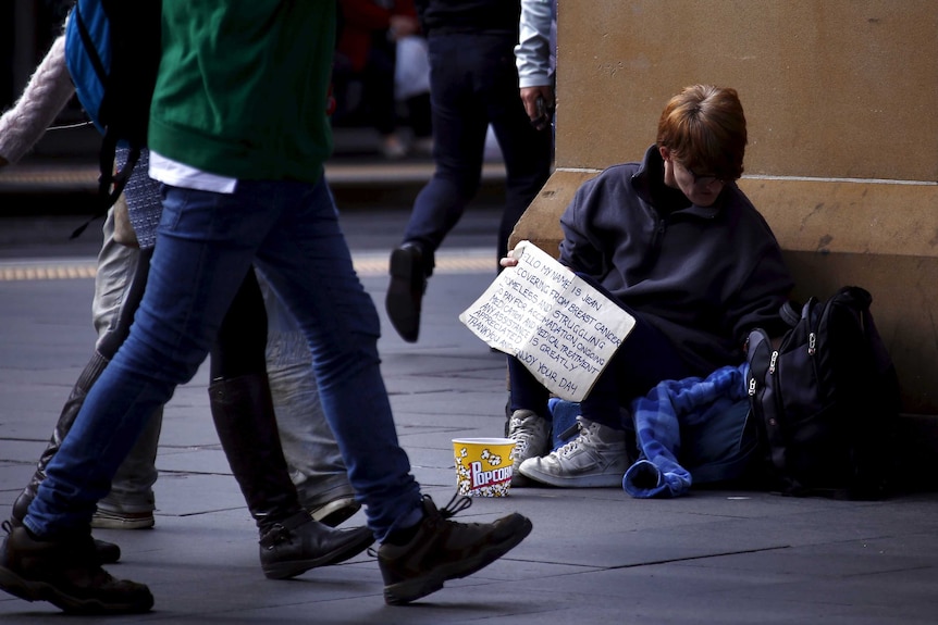 A woman holds a sign asking for money as pedestrians walk past