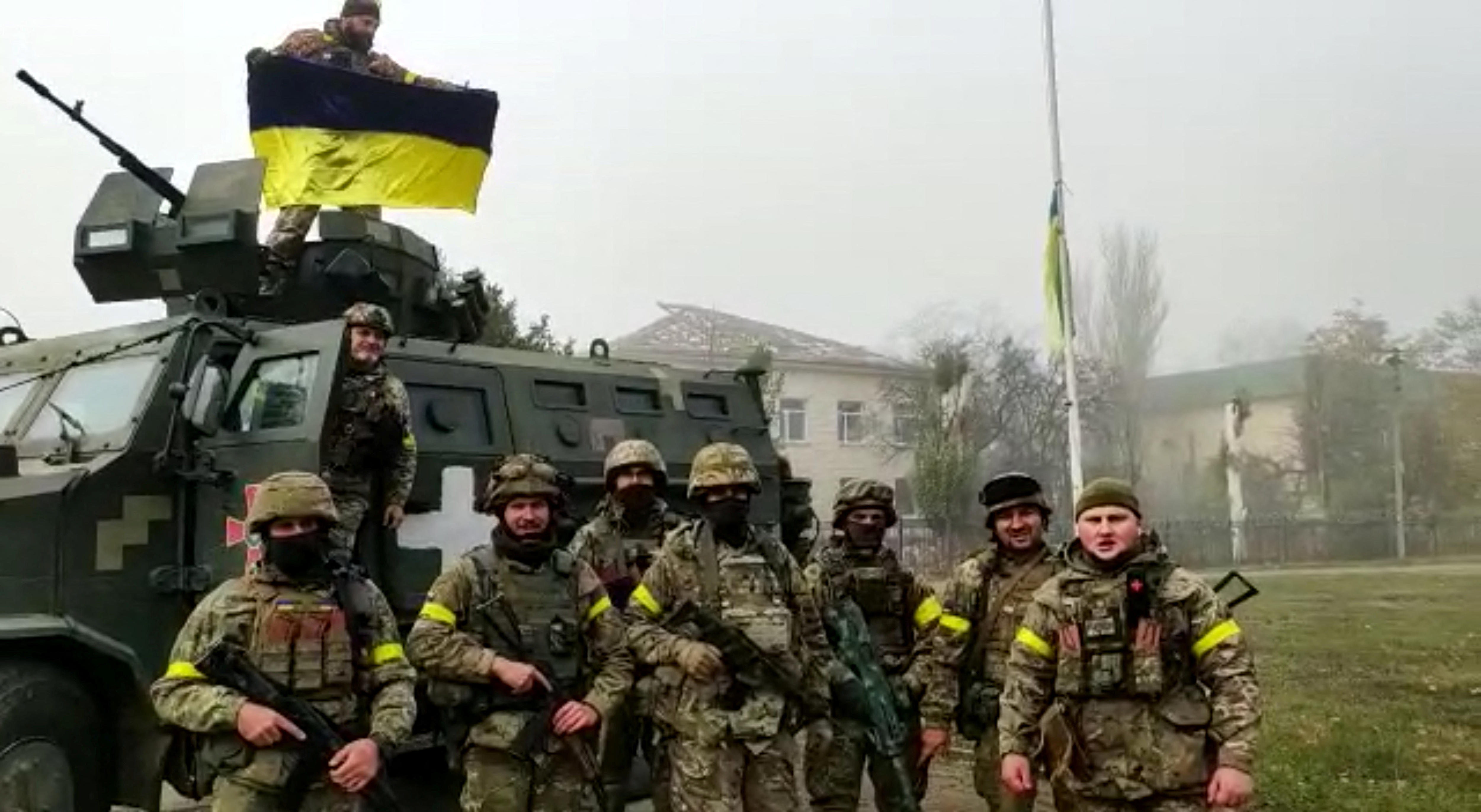 Soldiers pose for photo in front of vehicle, with Ukrainian flag hoisted on flagpole.