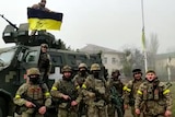 Soldiers pose for photo in front of vehicle, with Ukrainian flag hoisted on flagpole.